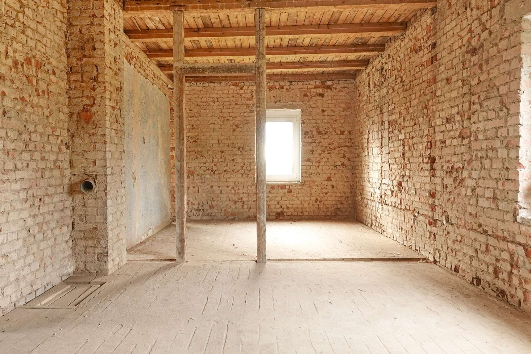 Unfinished brick interior with exposed walls, wooden ceiling beams, and two vertical support posts in a room lit by a small window at the far wall