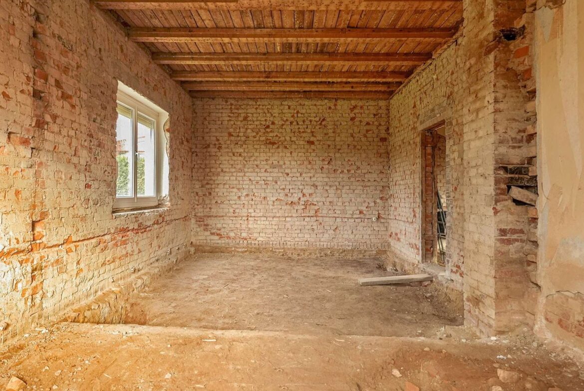 Unfinished brick room under renovation with exposed brick walls, a window on the left, and wooden ceiling beams overhead.