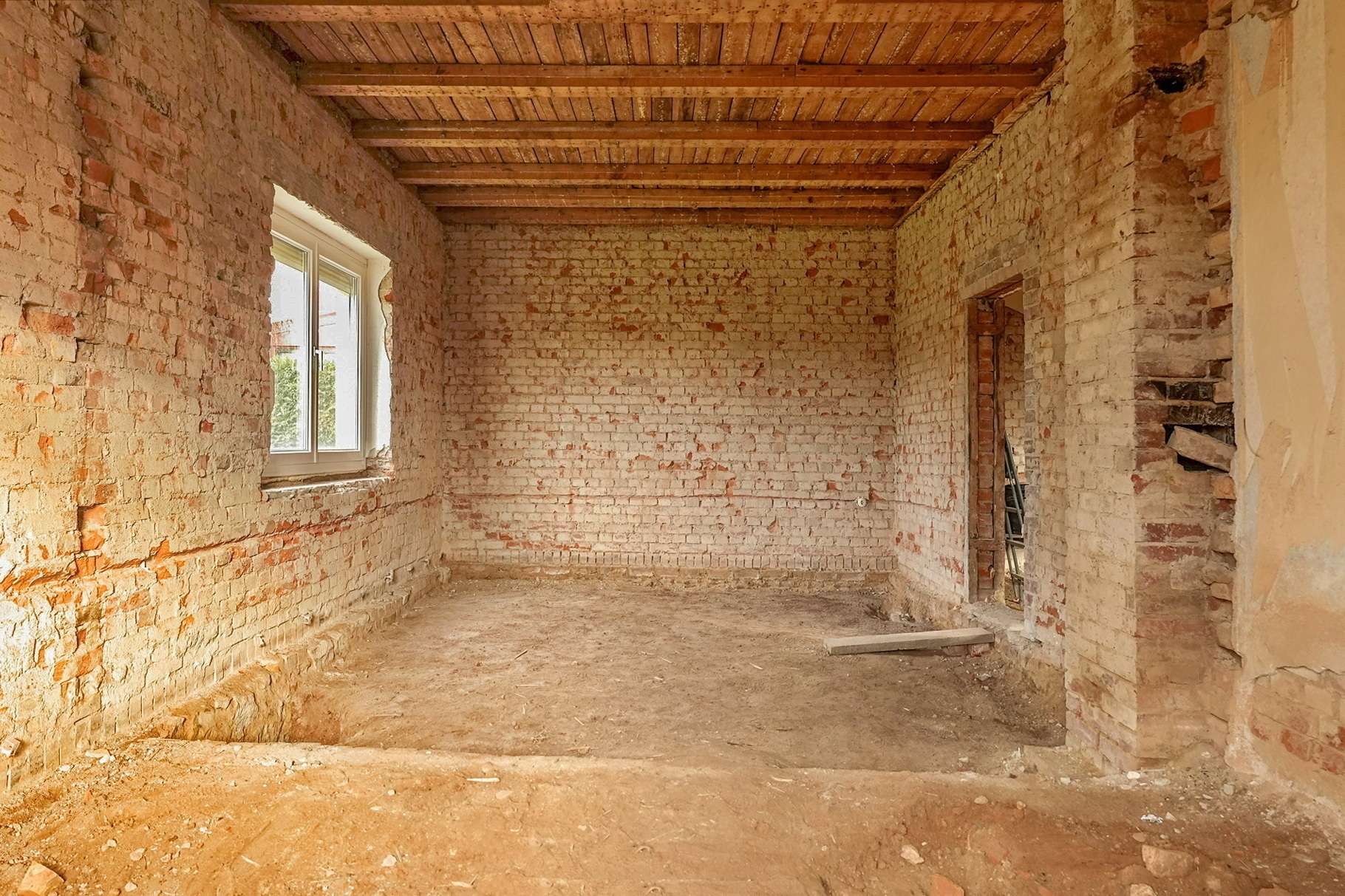 Unfinished brick room under renovation with exposed brick walls, a window on the left, and wooden ceiling beams overhead.
