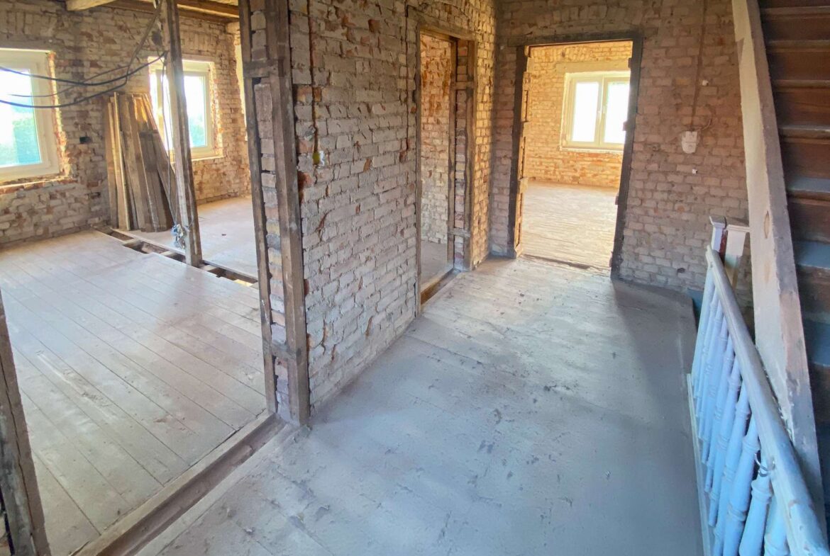 Interior of a brick-walled construction site with exposed beams and a doorway leading to a sunlit room.