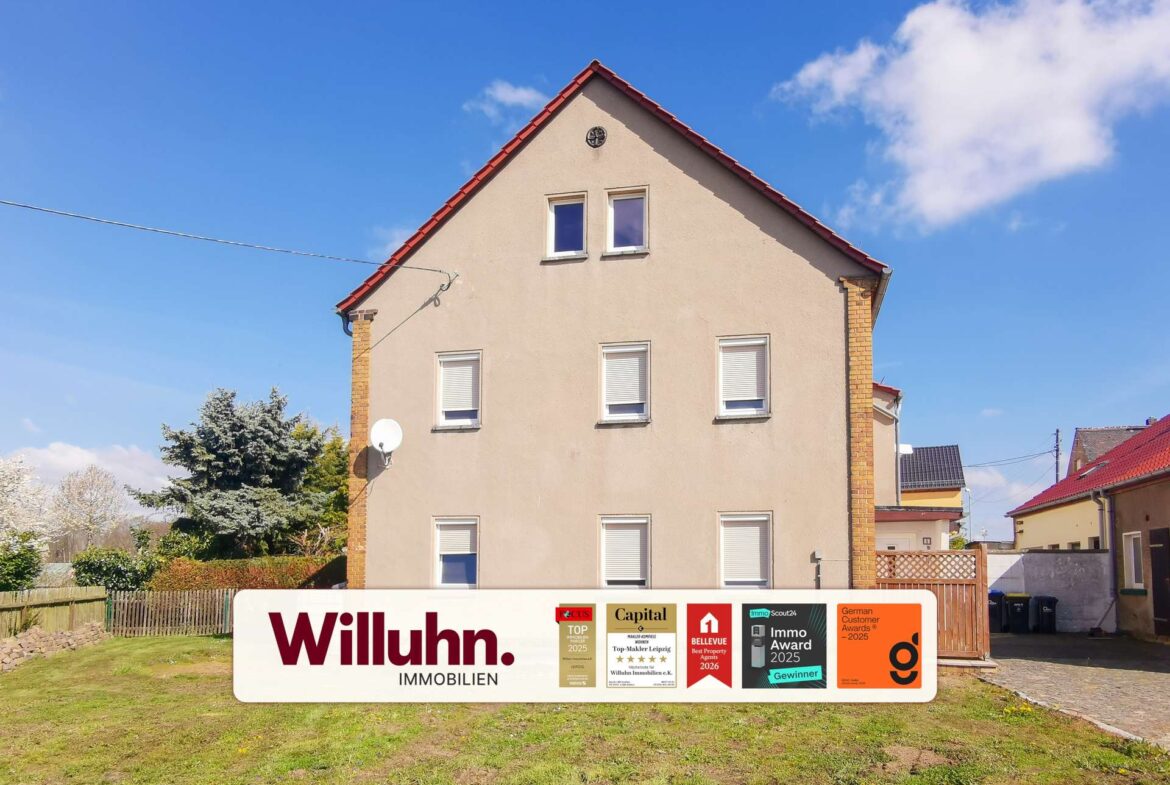 Beige multi-story house with a red gable roof under a blue sky; a large Willuhn Immobilien real estate sign with awards in the front yard.