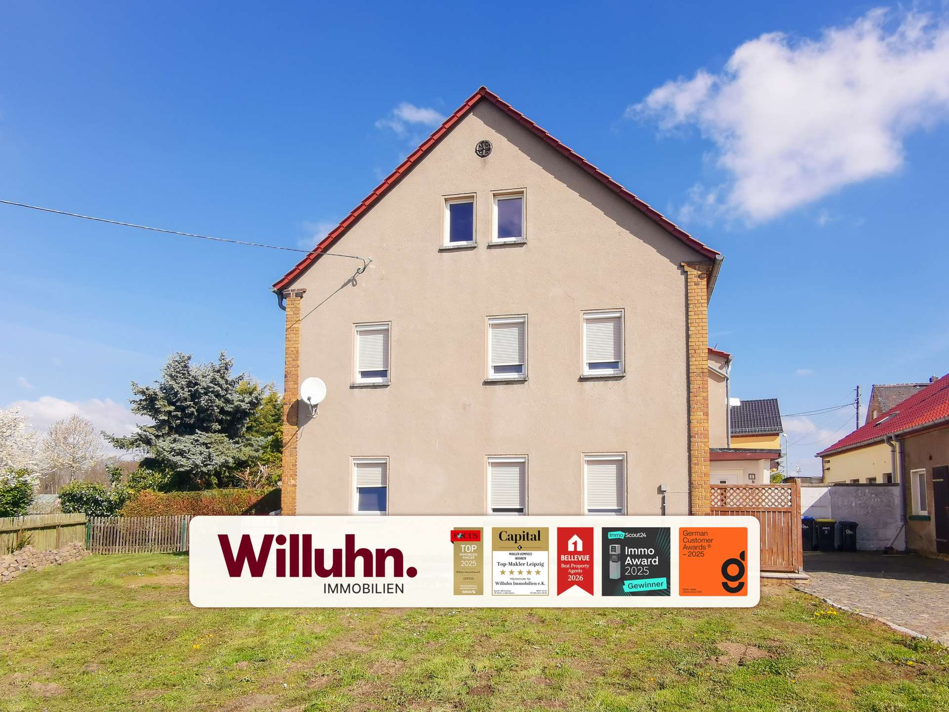 Beige multi-story house with a red gable roof under a blue sky; a large Willuhn Immobilien real estate sign with awards in the front yard.