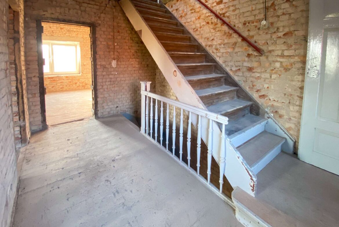 Interior of an unfinished brick stairwell with wooden stairs and white railing, exposed brick walls and a doorway to a sunlit room.