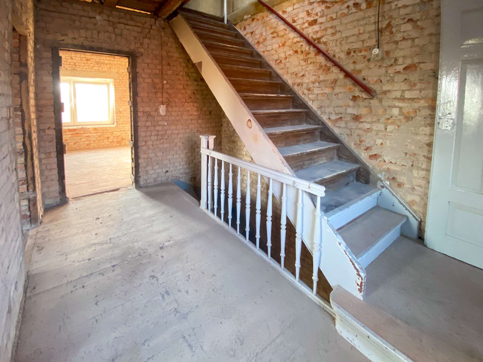 Interior of an unfinished brick stairwell with wooden stairs and white railing, exposed brick walls and a doorway to a sunlit room.