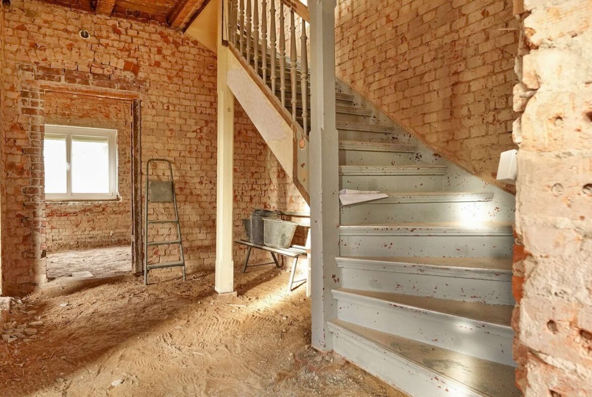 Unfinished brick-walled interior with a wooden staircase and a light blue metal staircase, construction debris on the dusty floor.