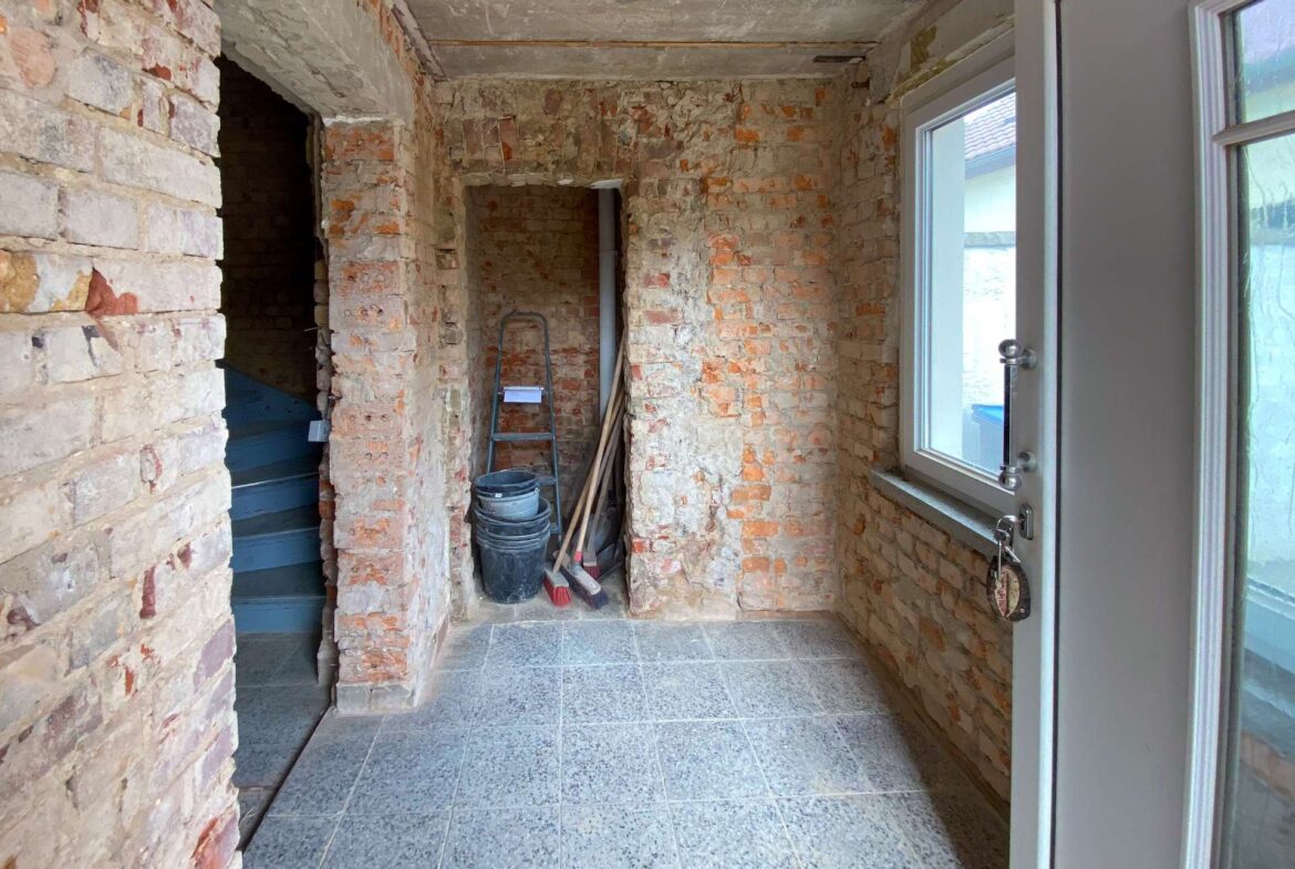 Interior of a partially finished brick-walled room with tools (ladder, buckets, broom) near a door with glass beside a windowed wall outside layout shows ongoing renovation.