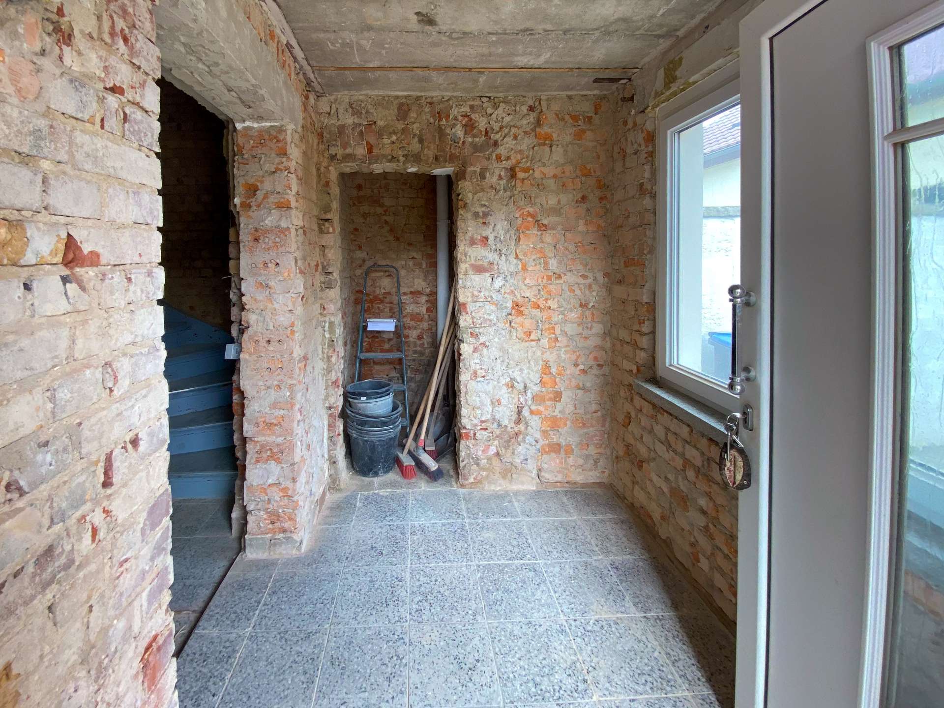 Interior of a partially finished brick-walled room with tools (ladder, buckets, broom) near a door with glass beside a windowed wall outside layout shows ongoing renovation.