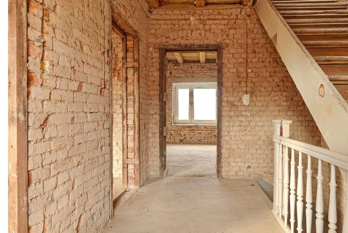 Unfinished brick hallway with exposed walls, a wooden staircase on the right, and a window at the far end letting in light.