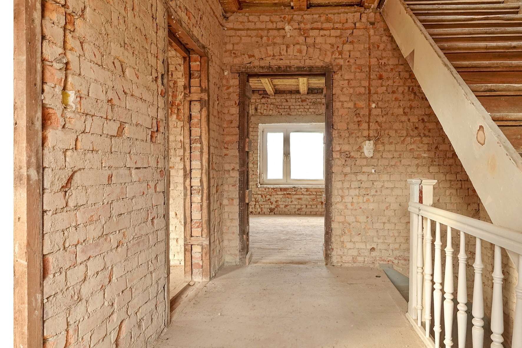 Unfinished brick hallway with exposed walls, a wooden staircase on the right, and a window at the far end letting in light.