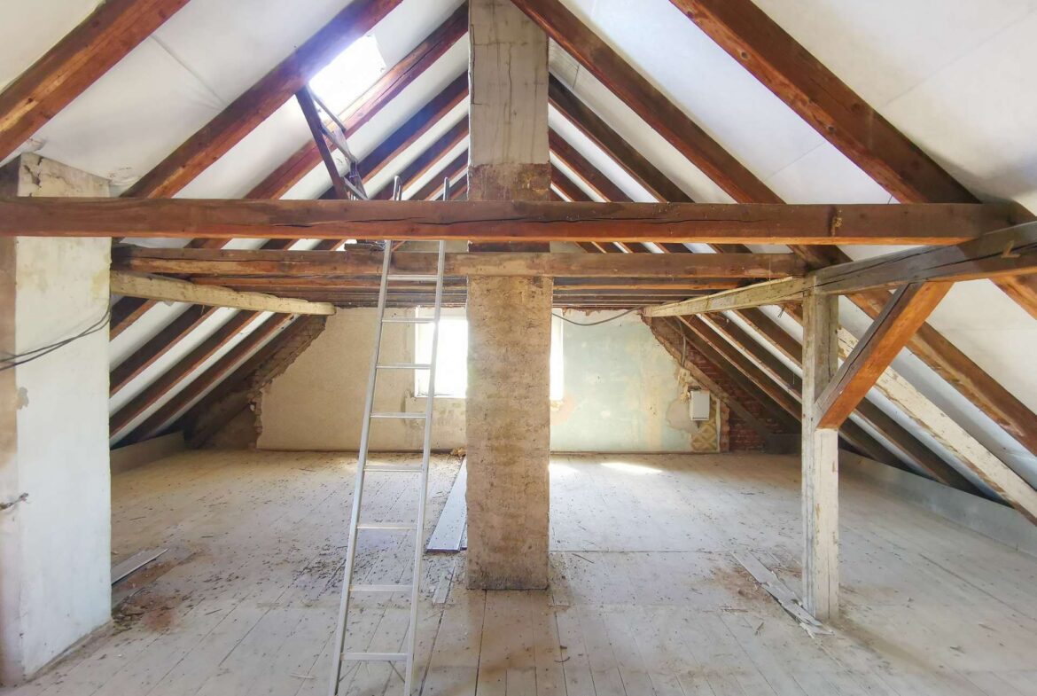 Attic with exposed wooden rafters, a metal ladder, and a central concrete pillar; peeling walls and dusty white floorboards reveal ongoing renovation.