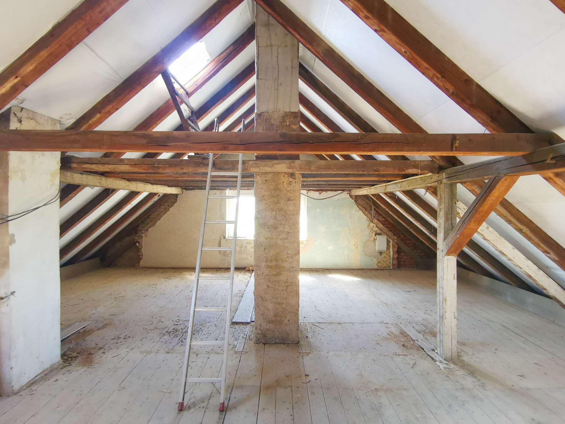 Attic with exposed wooden rafters, a metal ladder, and a central concrete pillar; peeling walls and dusty white floorboards reveal ongoing renovation.