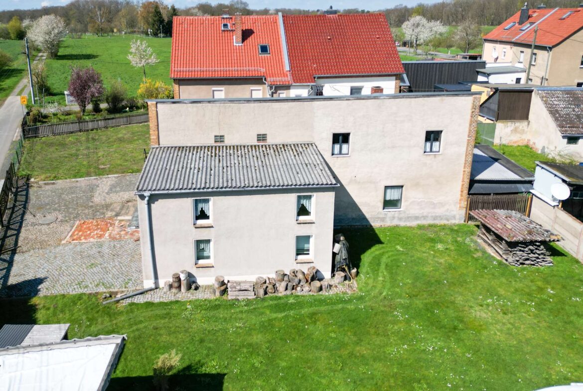 Aerial view of a backyard and houses: green lawn, stacked firewood, and red-tiled rooftops.