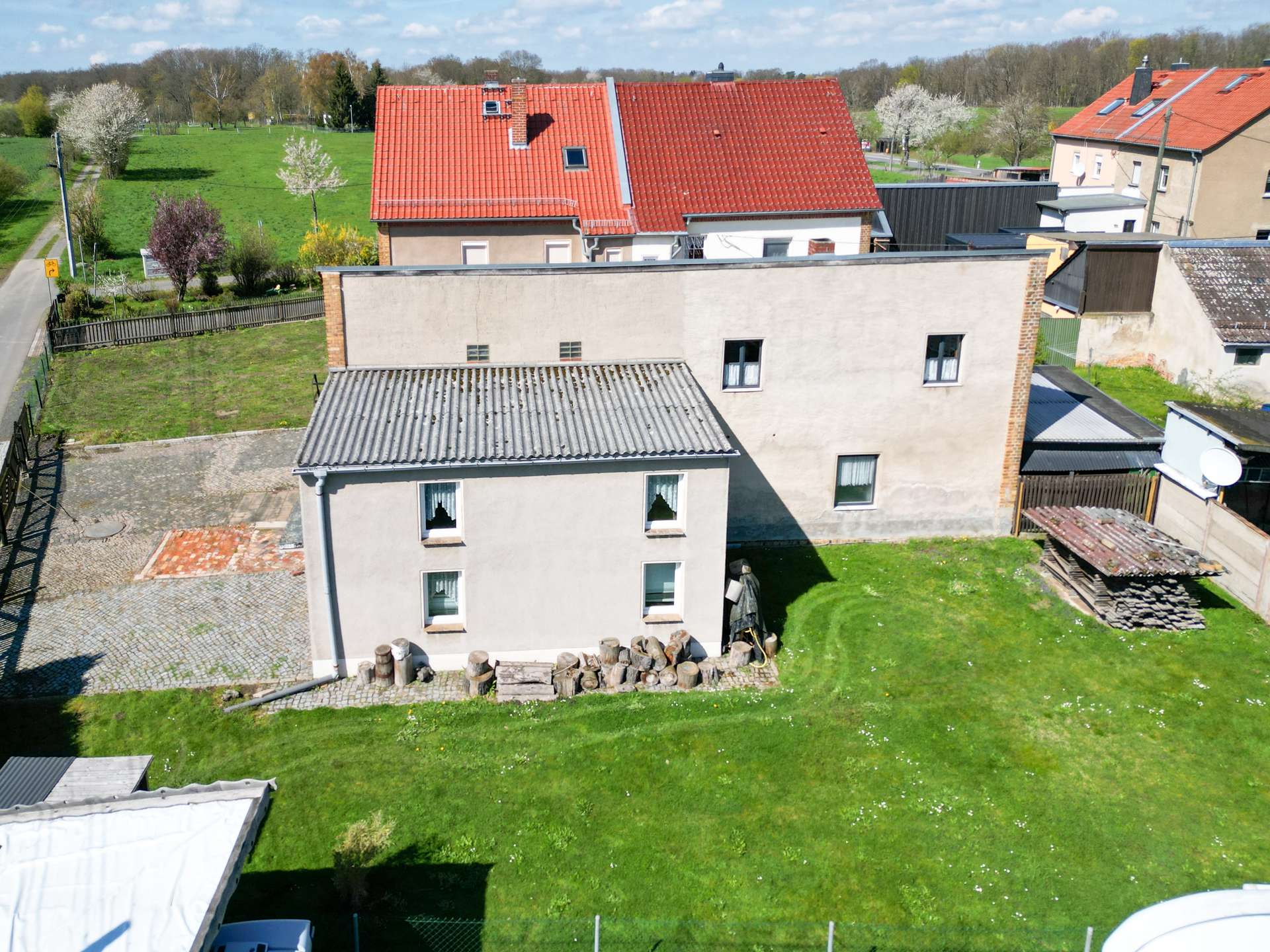 Aerial view of a backyard and houses: green lawn, stacked firewood, and red-tiled rooftops.
