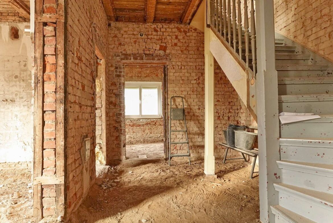 Unfinished brick interior under renovation, with a gray staircase on the right and a metal ladder in front of a window.