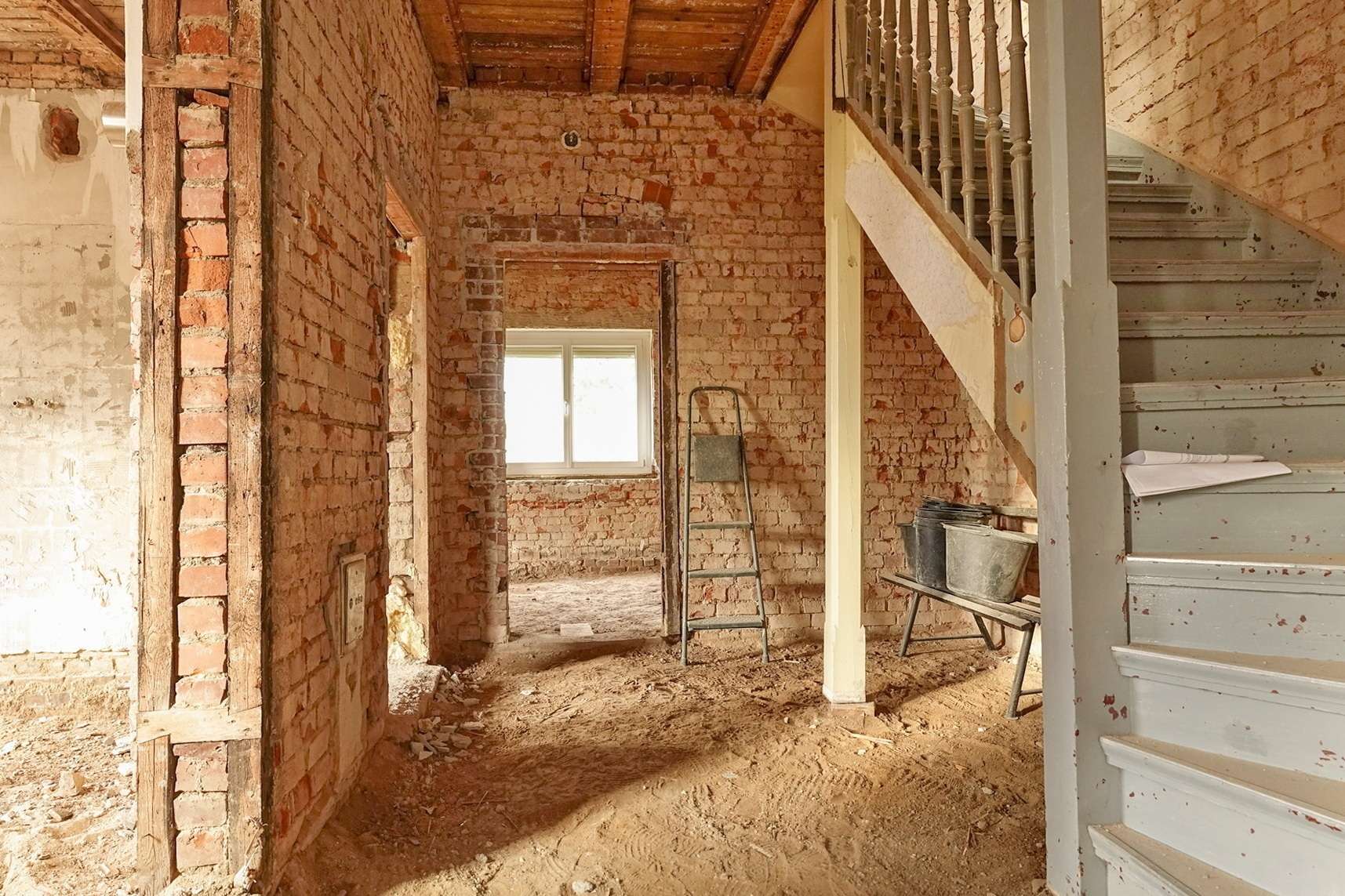Unfinished brick interior under renovation, with a gray staircase on the right and a metal ladder in front of a window.