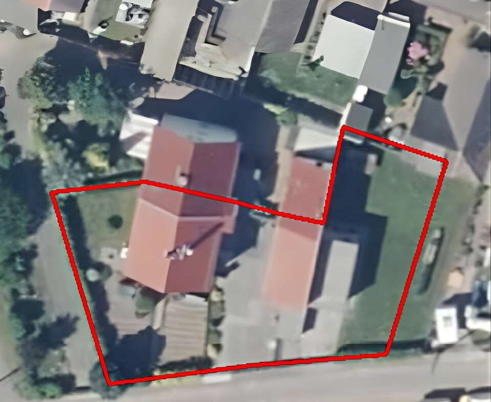 Aerial view of a residential area with a red boundary line outlining a lot that includes multiple buildings and a green lawn; a road runs along the bottom, with neighboring houses and trees visible, and a compass rose appears in the top-right corner.