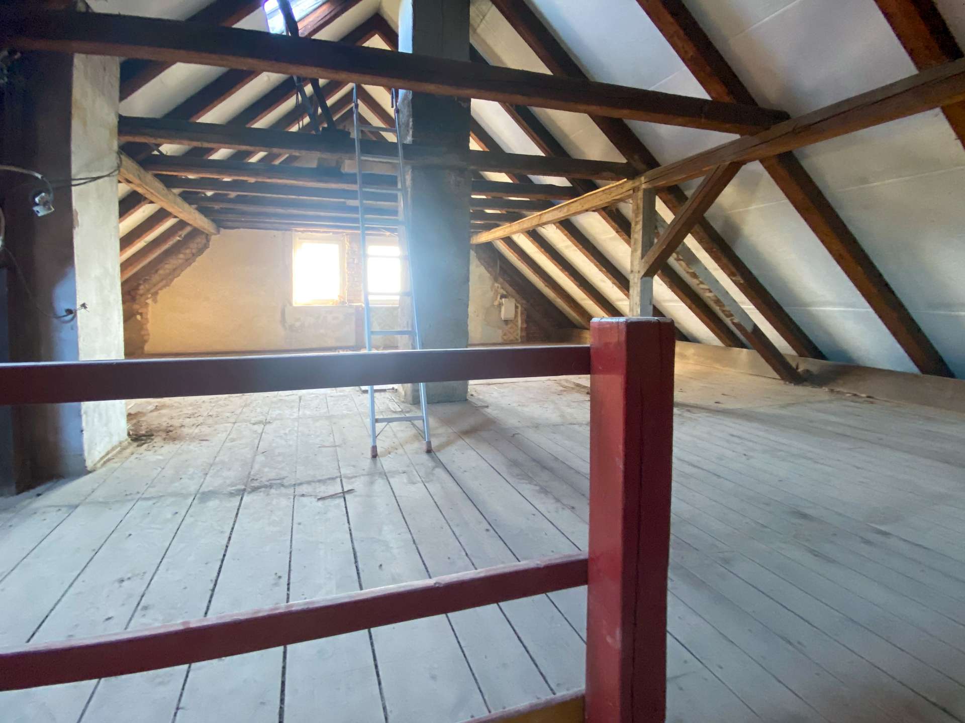 Dusty attic with exposed wooden beams, a red railing in the foreground, and a ladder by a bright window.