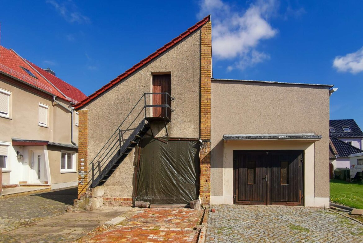 Beige residential building with a red sloped roof, brick chimney, and an exterior metal staircase leading to a wooden door on the upper level under a blue sky.