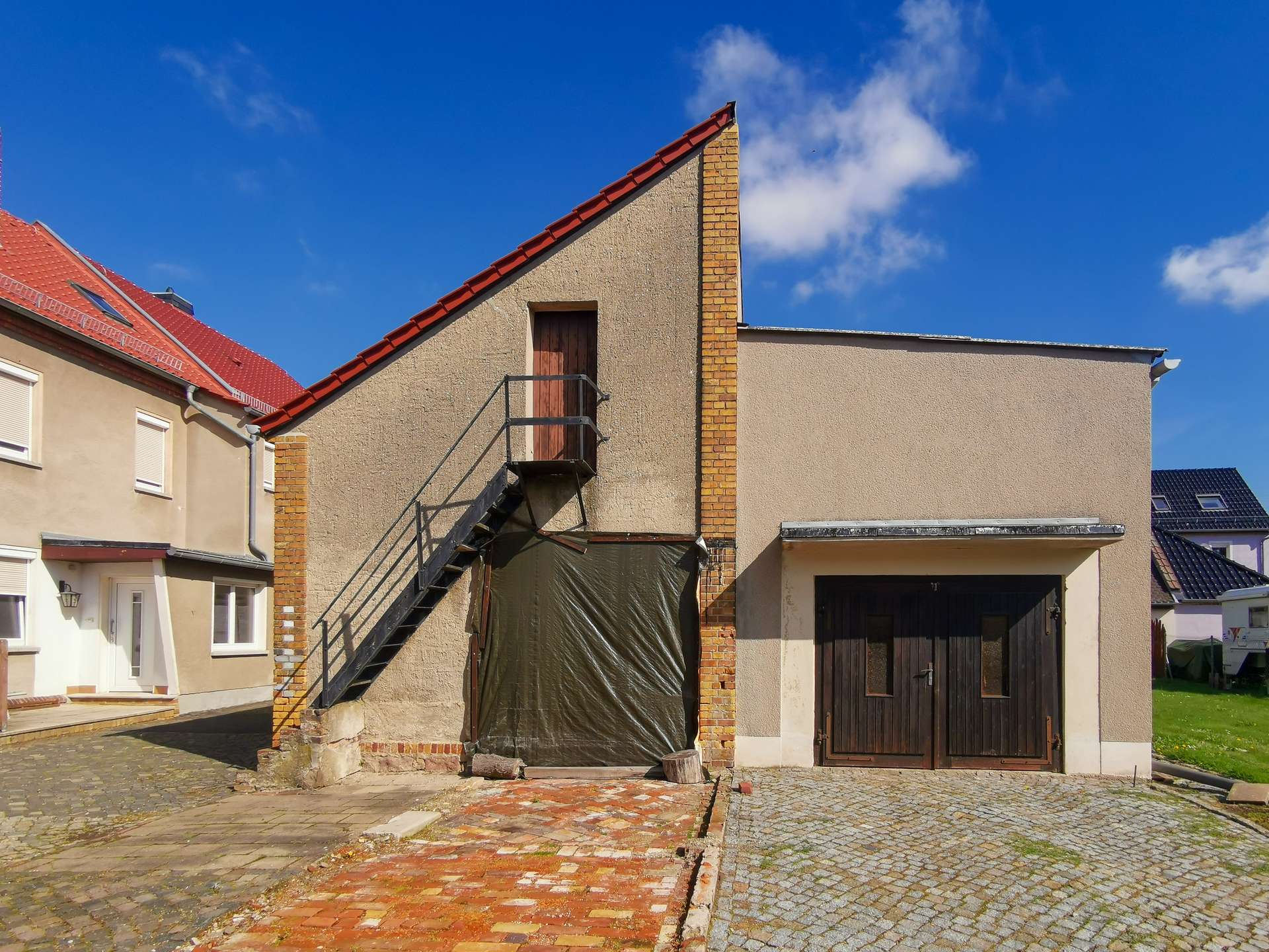 Beige residential building with a red sloped roof, brick chimney, and an exterior metal staircase leading to a wooden door on the upper level under a blue sky.