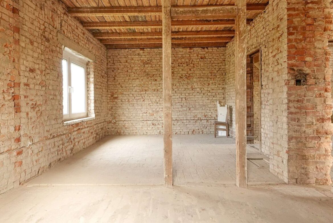 Interior of an unfinished brick-walled room with wooden ceiling beams, a window on the left, and a chair against the far wall.