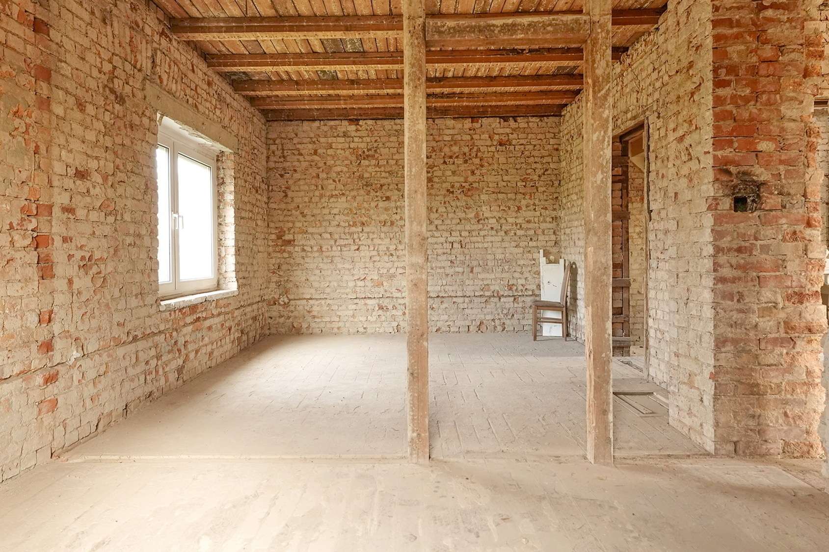 Interior of an unfinished brick-walled room with wooden ceiling beams, a window on the left, and a chair against the far wall.
