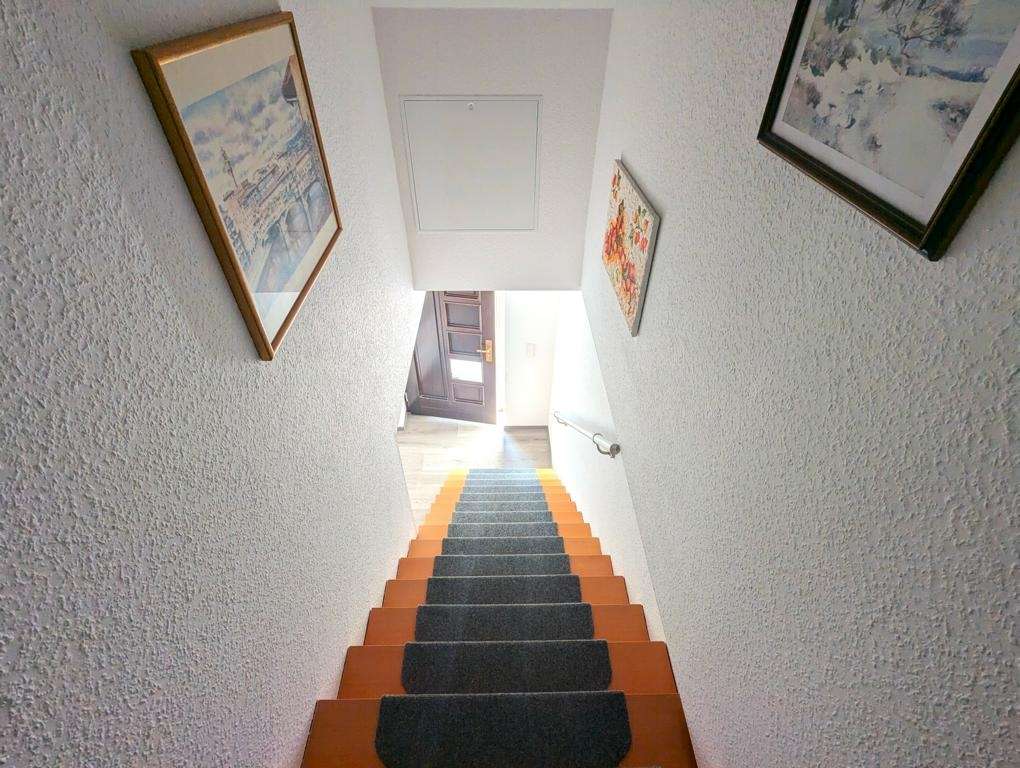 Narrow interior staircase viewed from above, orange and gray carpet runners on each step, white textured walls, framed pictures, and a door at the bottom with a railing on the right side.