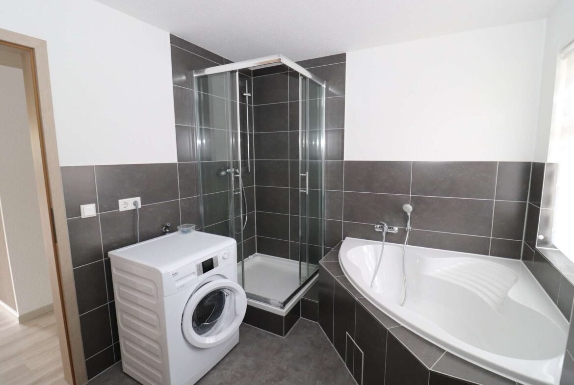 Modern bathroom with a corner glass shower enclosure, a freestanding bathtub, and a white front-loading washing machine against dark gray tiled walls.