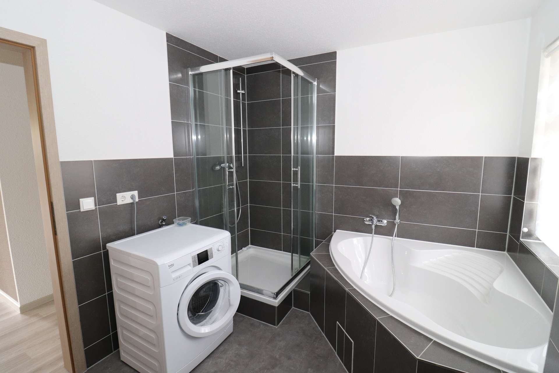 Modern bathroom with a corner glass shower enclosure, a freestanding bathtub, and a white front-loading washing machine against dark gray tiled walls.