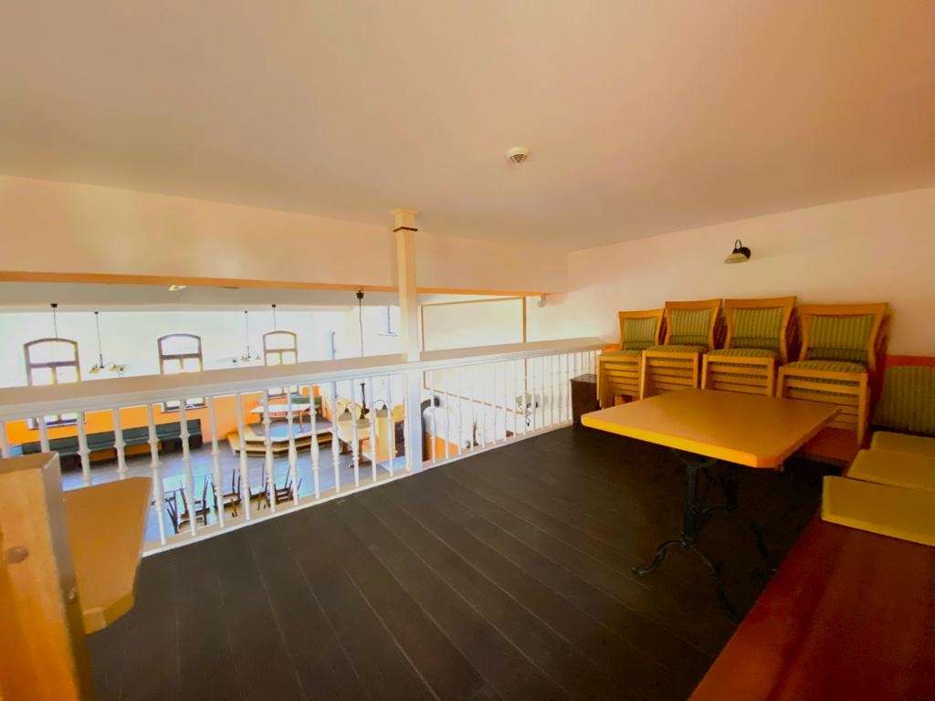 Loft lounge area with a wooden table and green-cushioned chairs along the wall, overlooking a lower dining space behind a white railing.