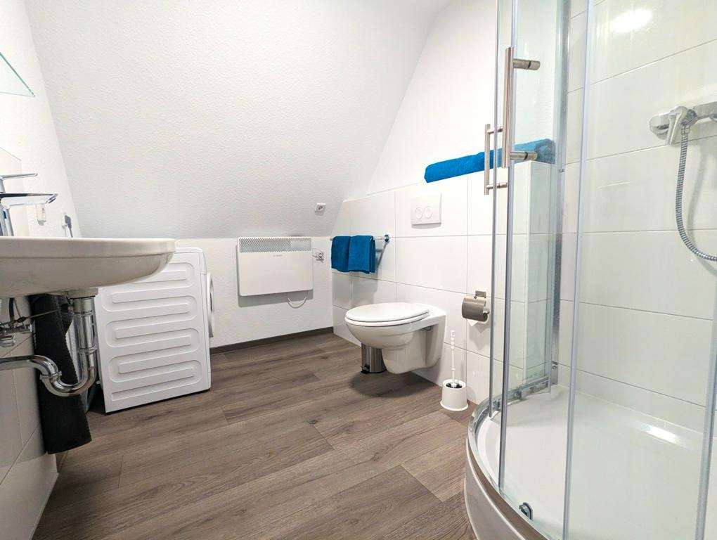 Bright bathroom with a curved ceiling, glass corner shower, wall-mounted toilet, blue towels, sink, and a front-loading washing machine on the left.