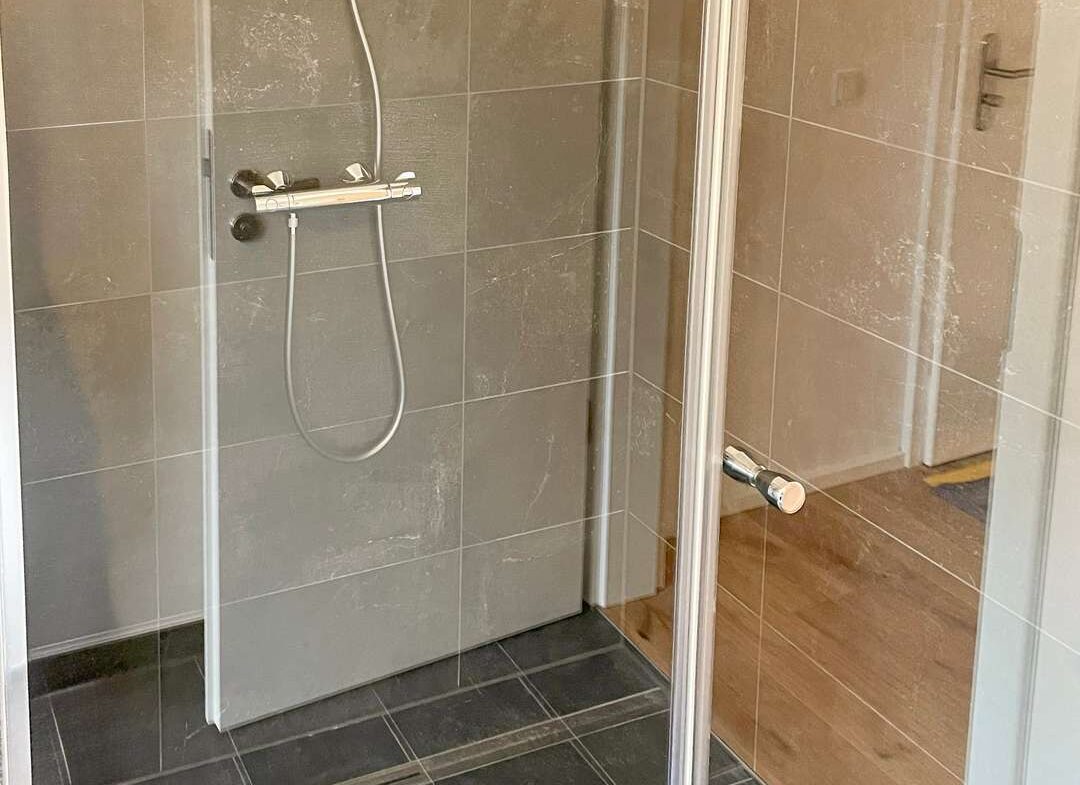 Shower enclosure with a handheld shower on a rail, gray tiled walls, and a glass door.