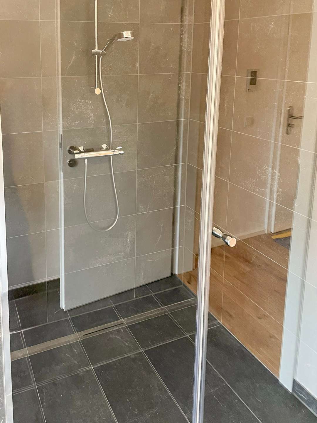 Shower enclosure with a handheld shower on a rail, gray tiled walls, and a glass door.