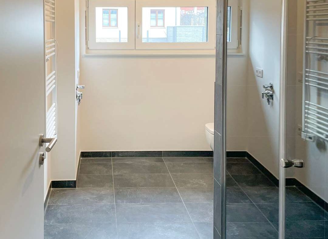 Modern bathroom with gray floor tiles, glass shower enclosure on the right, white walls, a wall-mounted toilet, and a towel radiator near the door.