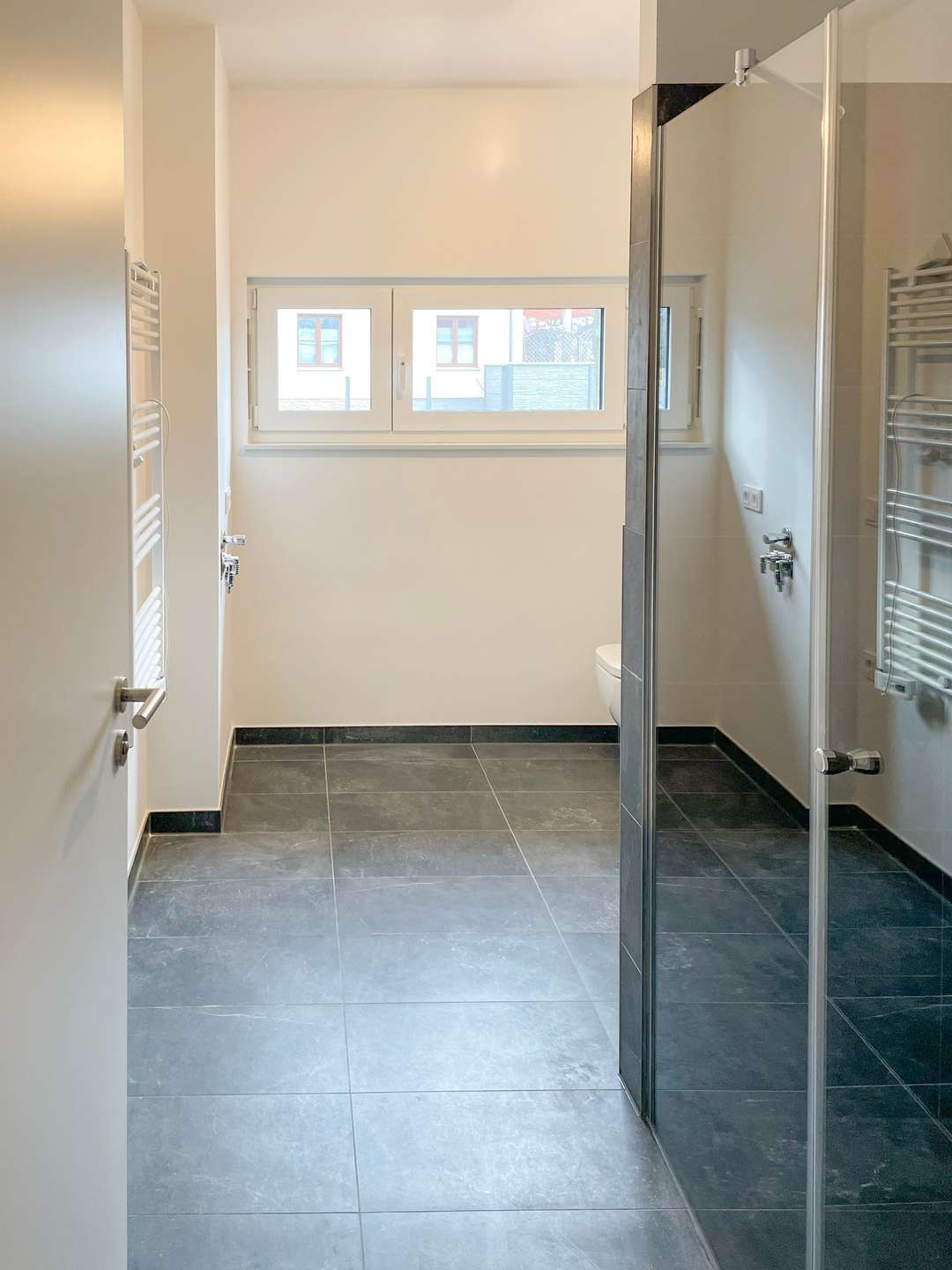 Modern bathroom with gray floor tiles, glass shower enclosure on the right, white walls, a wall-mounted toilet, and a towel radiator near the door.