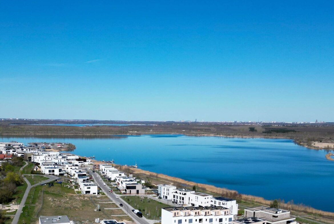 Aerial view of a modern residential neighborhood along a wide river, white apartment buildings lining streets under a clear blue sky.