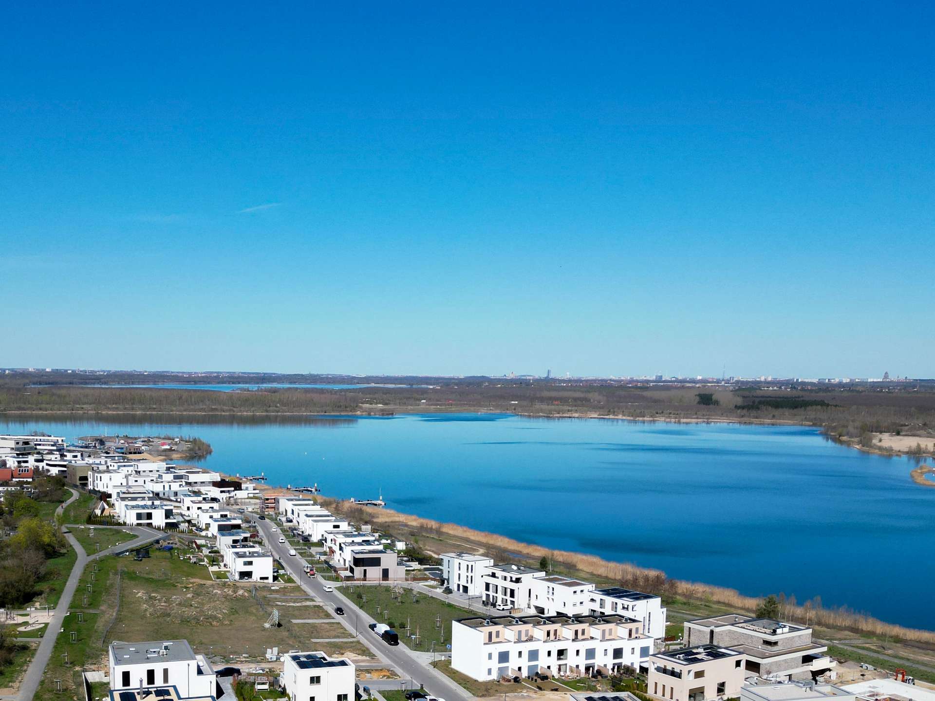 Aerial view of a modern residential neighborhood along a wide river, white apartment buildings lining streets under a clear blue sky.