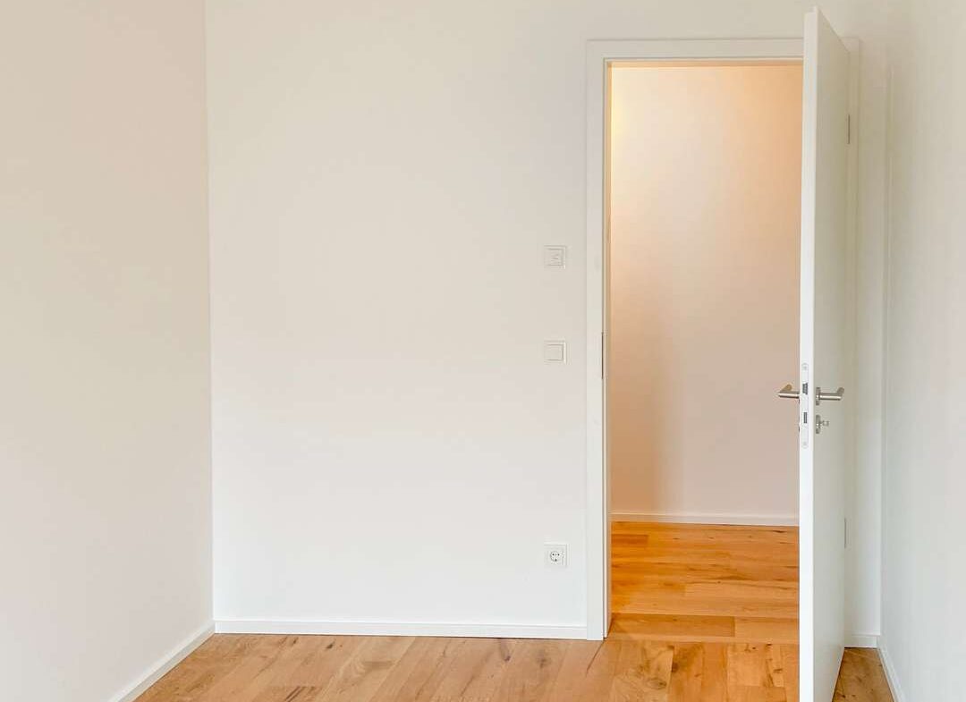 Empty white-walled room with light wood floor and an open white door leading to a bright adjacent room.