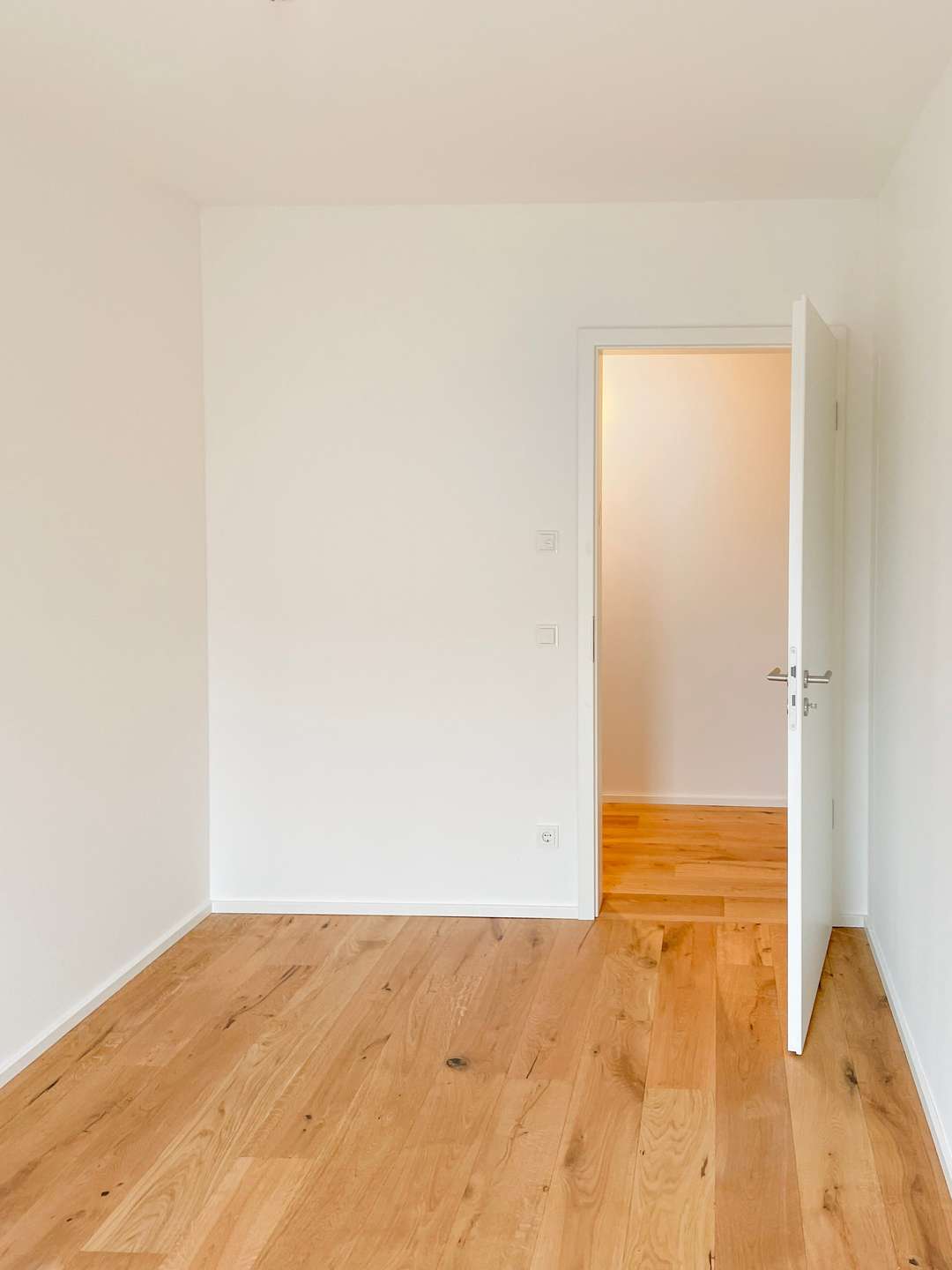 Empty white-walled room with light wood floor and an open white door leading to a bright adjacent room.