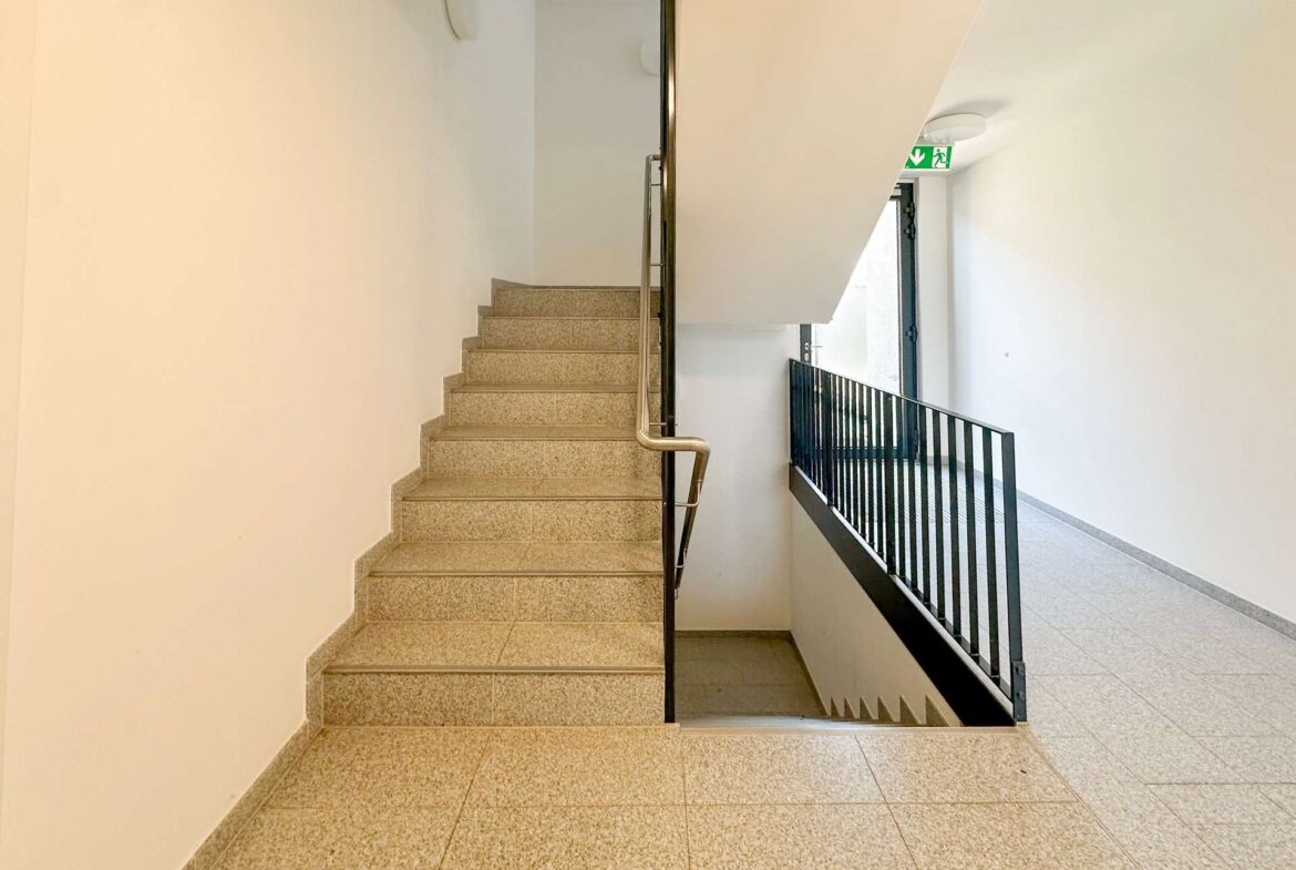 Interior stairwell with beige tiled steps, metal handrail, and a railing along the level; exit door and green emergency exit sign visible in the distance.