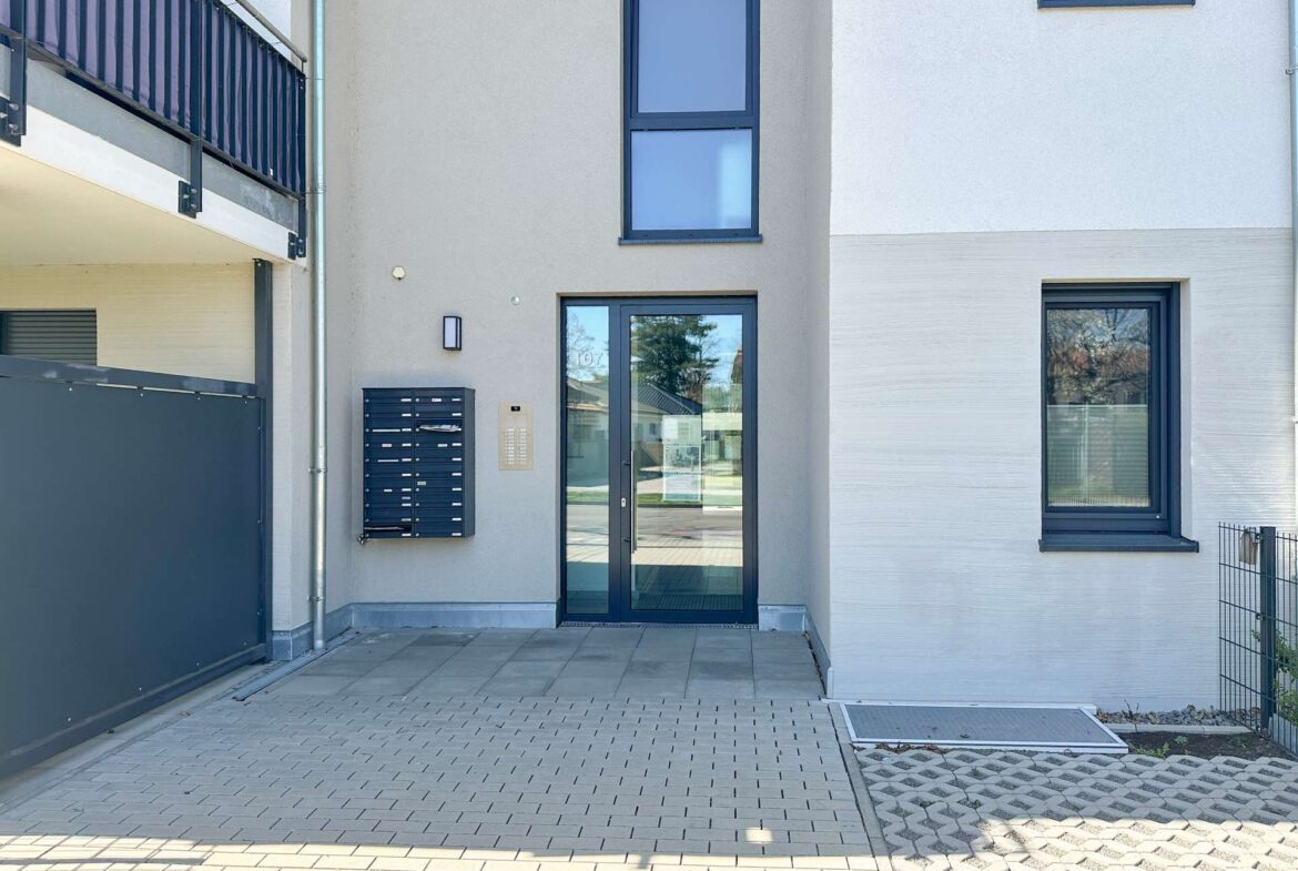 Modern apartment building entrance with glass doors, gray walls, blue-framed windows, and mailboxes beside an intercom panel.