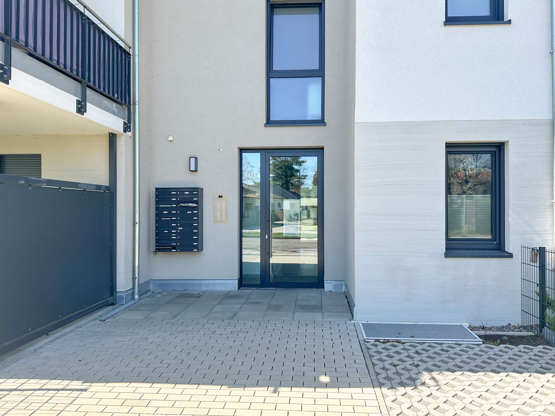 Modern apartment building entrance with glass doors, gray walls, blue-framed windows, and mailboxes beside an intercom panel.