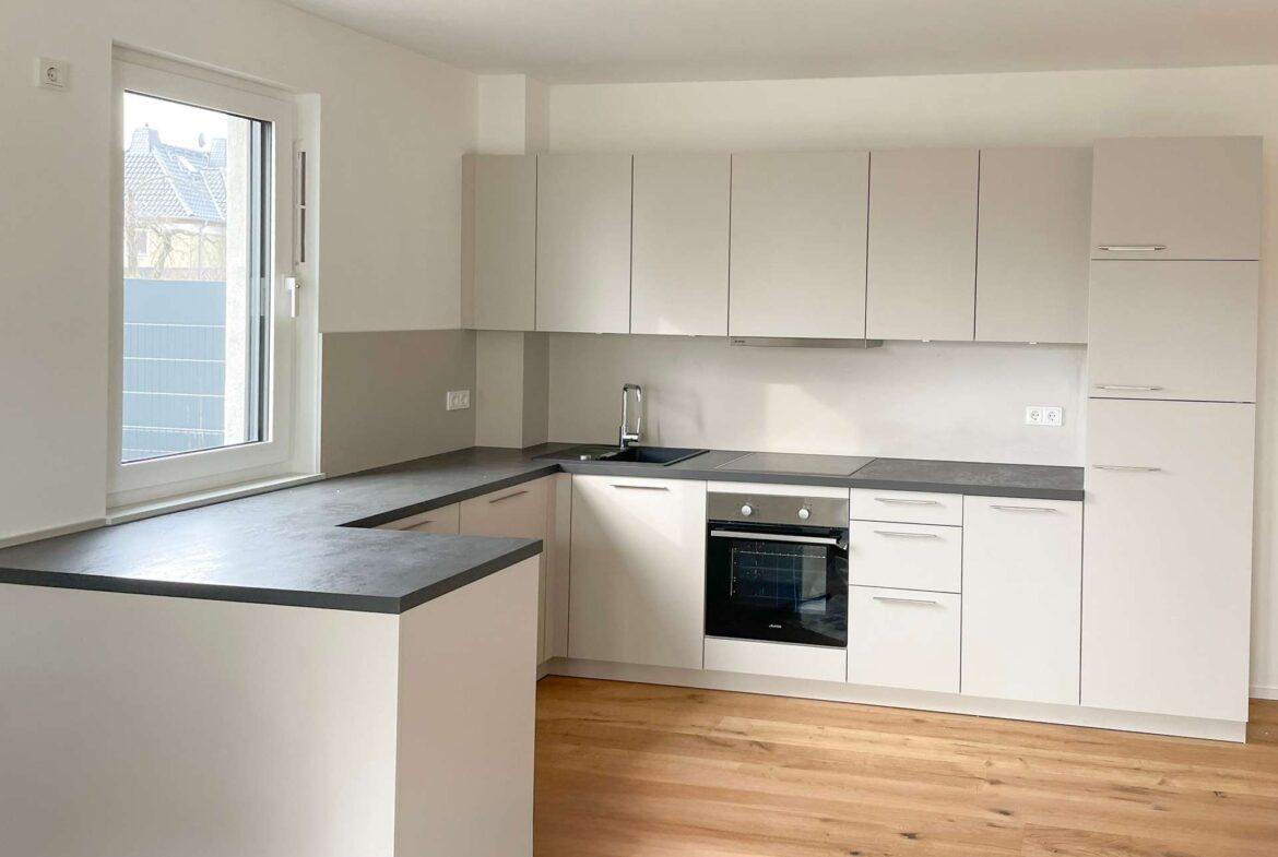Modern U-shaped kitchen with white cabinets, grey countertops, and a built-in oven along a grey backsplash wall, wooden floor visible.