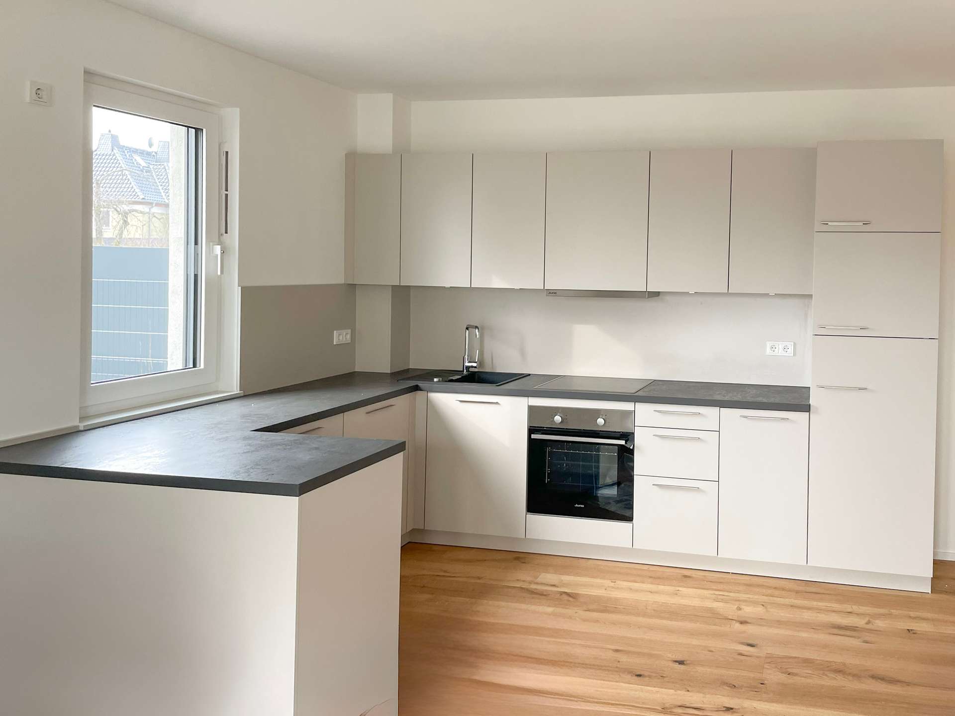 Modern U-shaped kitchen with white cabinets, grey countertops, and a built-in oven along a grey backsplash wall, wooden floor visible.