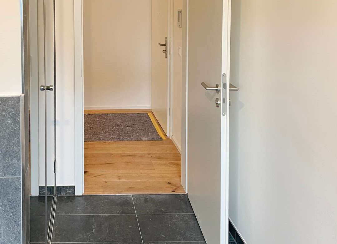 Entryway photo showing a transition from dark tiled floor to light wooden flooring, with an open door leading to a small hallway and a dark gray rug at the far end