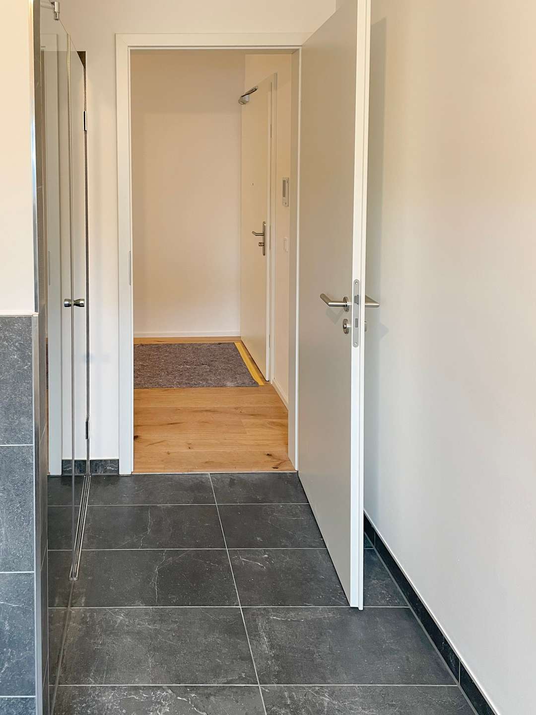 Entryway photo showing a transition from dark tiled floor to light wooden flooring, with an open door leading to a small hallway and a dark gray rug at the far end