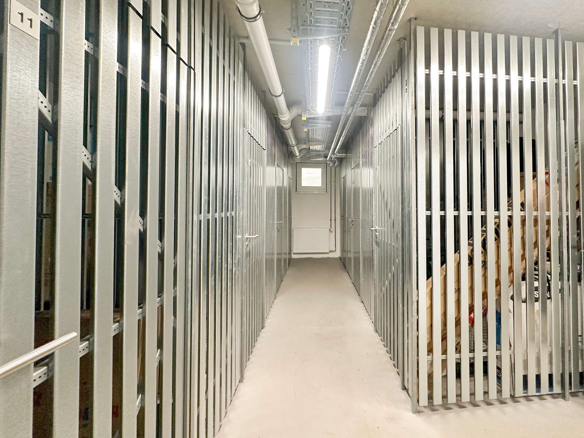 Narrow hallway between tall metal storage racks in a warehouse, with pipes and a fluorescent light overhead.