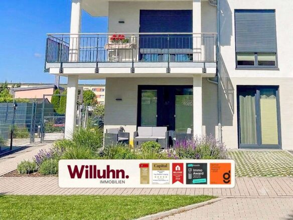 Modern two-story house with balcony, glass railing, patio seating, and a landscaped front yard; a Willuhn Immobilien sign with awards in the foreground.