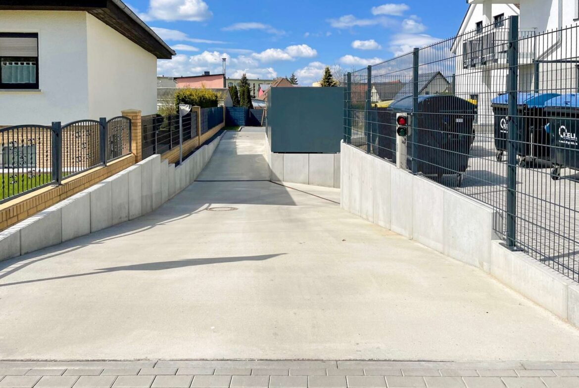 Wide residential driveway flanked by metal fences and concrete walls, with white houses and a blue sky; black bins behind the fence on the right.