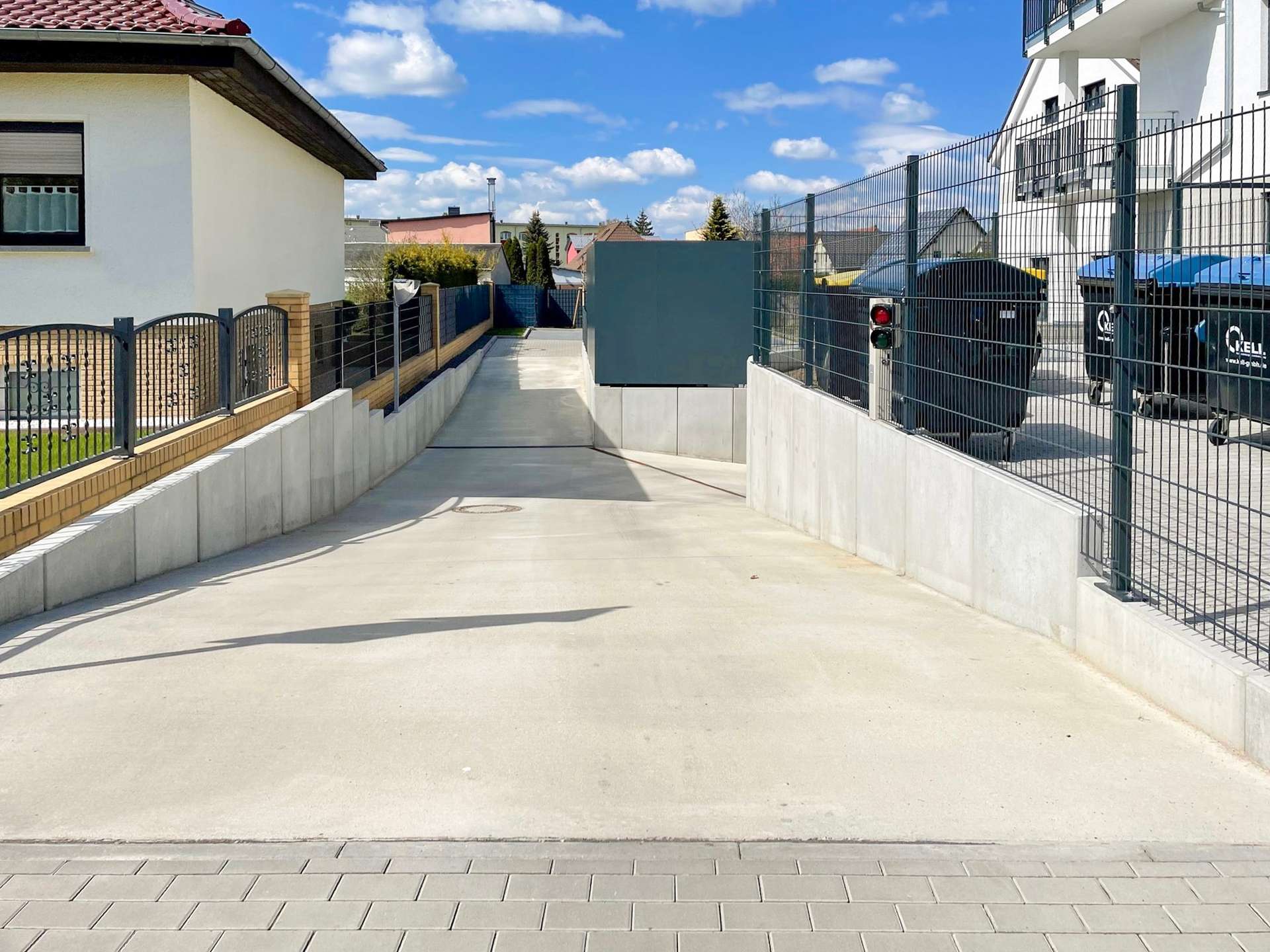 Wide residential driveway flanked by metal fences and concrete walls, with white houses and a blue sky; black bins behind the fence on the right.