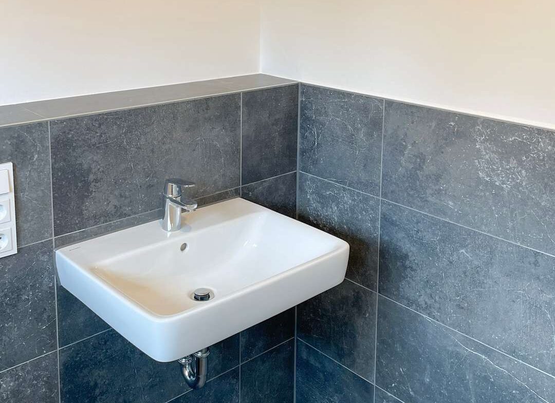 White wall-mounted sink with chrome faucet in a corner bathroom with dark gray tiled walls and floor.